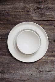 Overhead view of the Looks Like White | Scandi Cereal Bowl nested in a dinner plate on a rustic wooden table, highlighting elegance and simplicity.